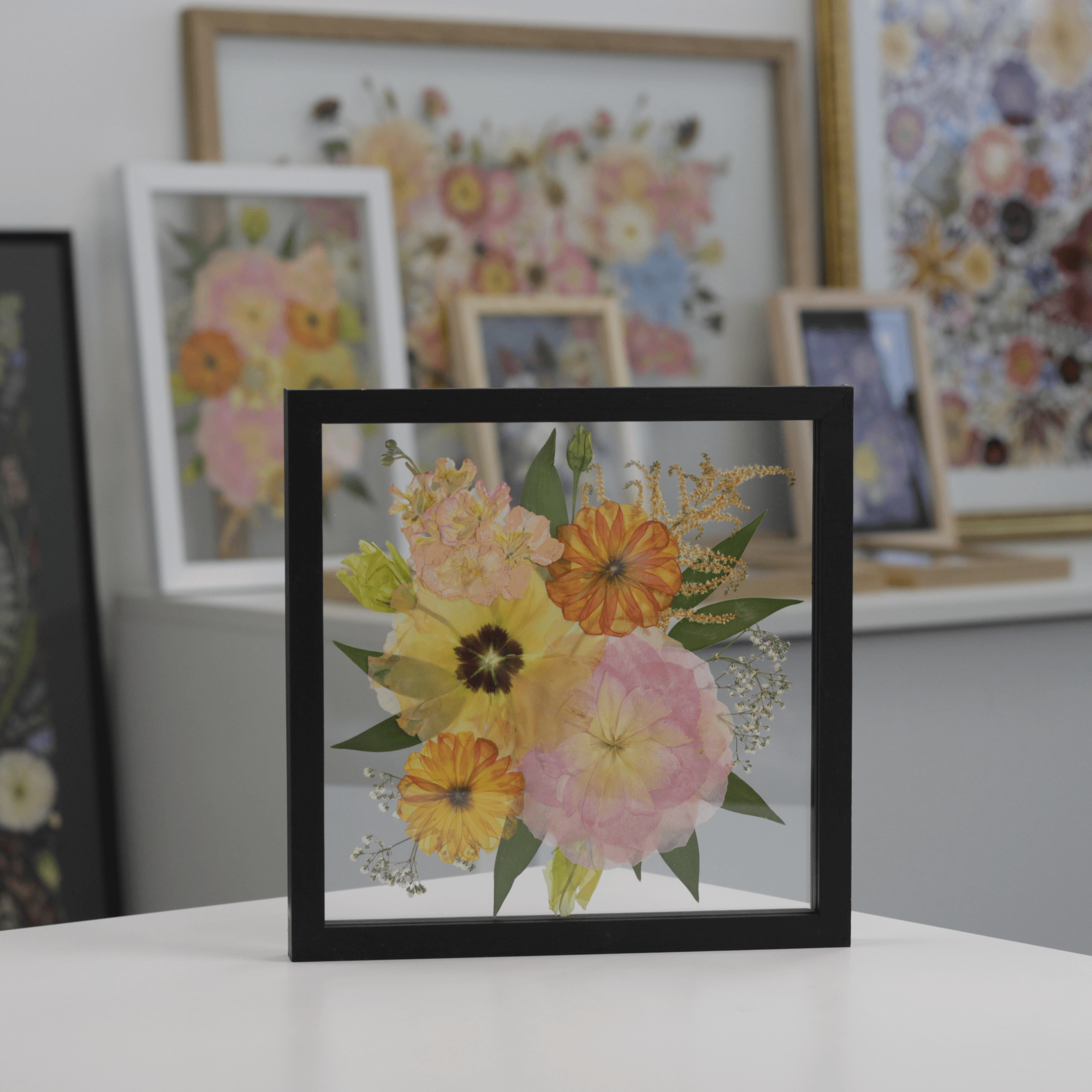 Framed pressed flower artwork in a black float frame displayed in front of more floral artworks in various frames