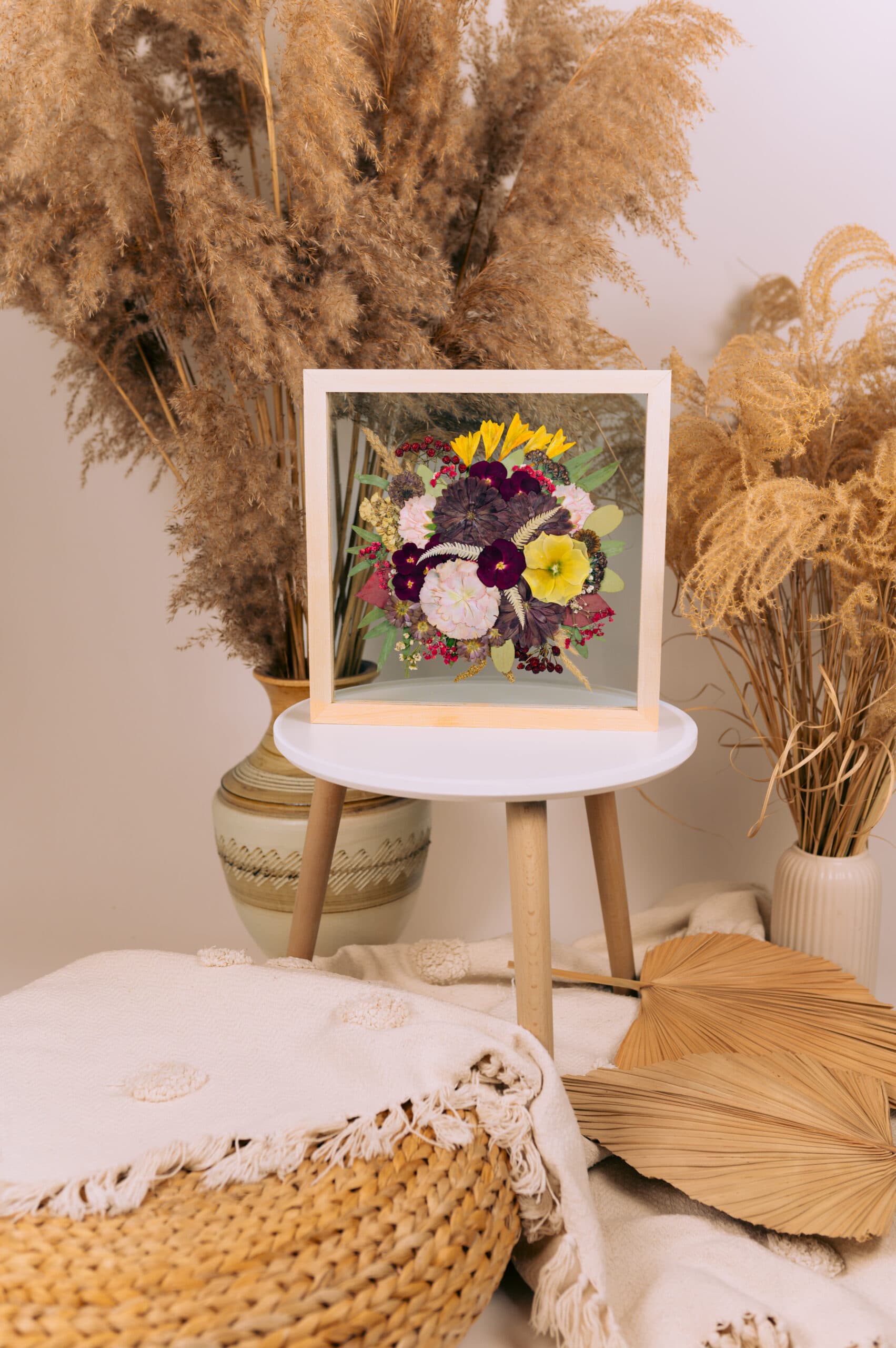 Framed pressed flower artwork displayed on a small side table surrounded by pampas grass and boho decor